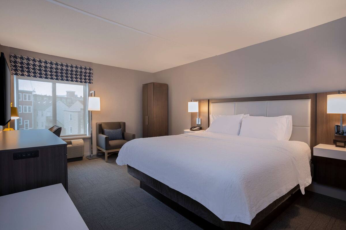 A hotel room with a large bed, a desk, a chair, a wardrobe, and a window with a view of buildings.