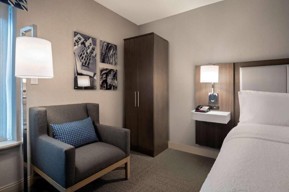 A hotel room with a bed, a chair, a wardrobe, a bedside table with a lamp, and wall art.
