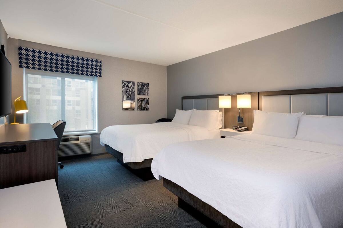A hotel room with two beds, a desk, a chair, a window, and a wall with pictures.