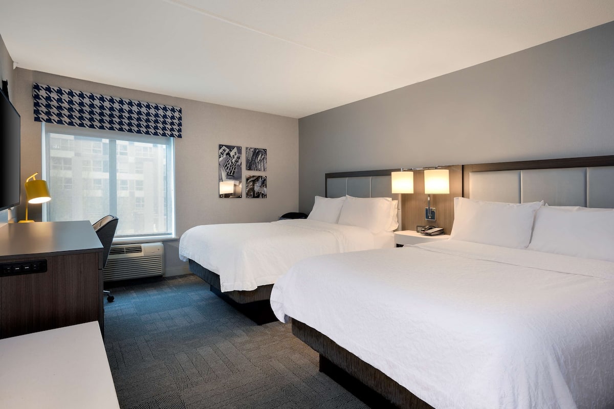 A hotel room with two beds, a desk, a chair, a desk lamp, and a window with a view of buildings.