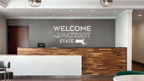 A reception area with a wooden reception desk, a sign reading "WELCOME TO THE PATRIOT STATE," and a map of the state on the wall. - Hampton Inn by Hilton Boston/Cambridge (Cambridge)