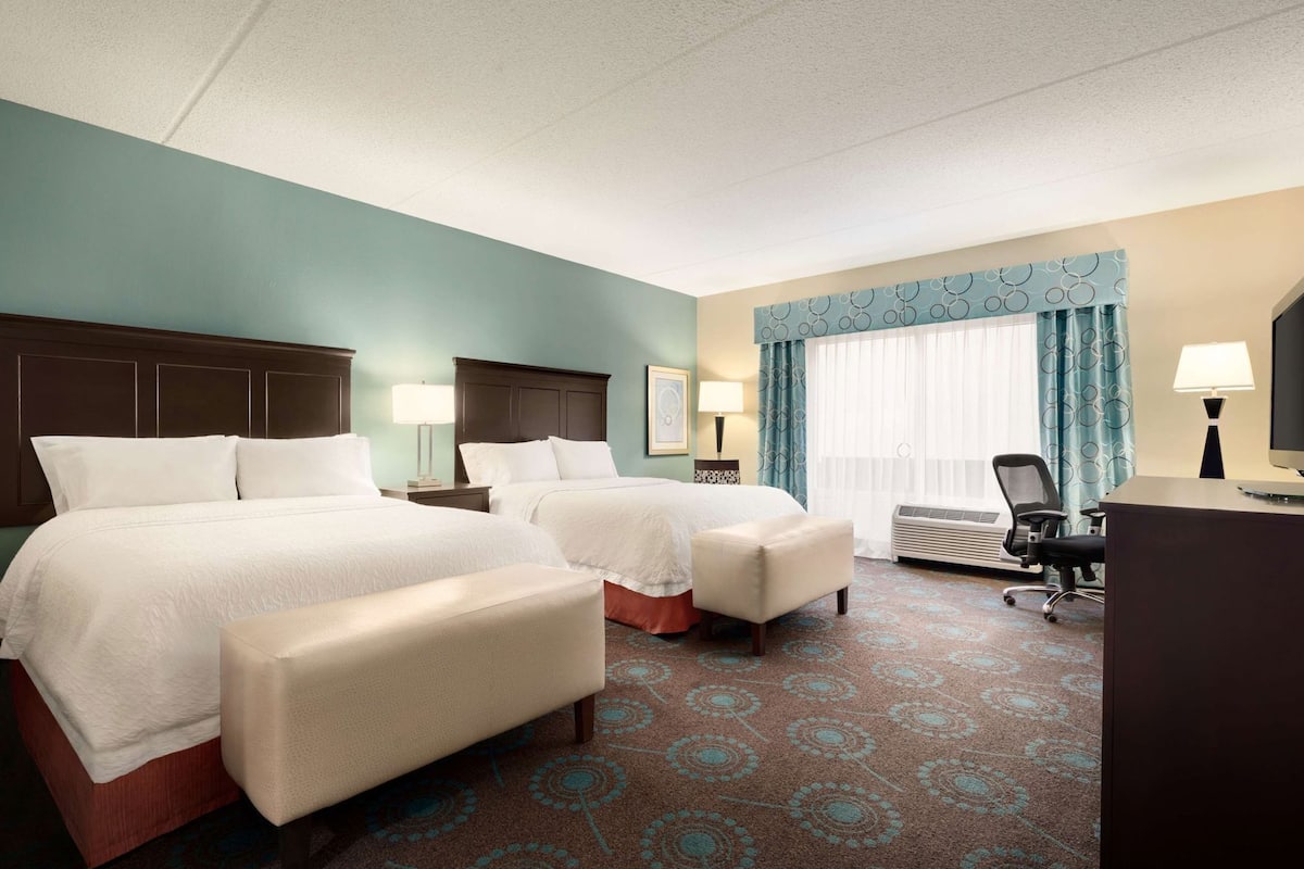 A hotel room with two beds, a desk, a chair, a window with curtains, and a patterned carpet.
