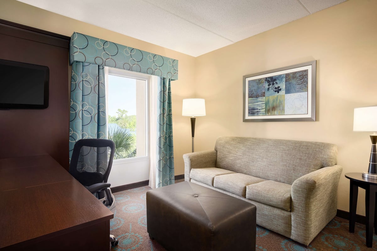 A hotel room with a wooden desk, a sofa, an ottoman, a chair, a lamp, and a painting on the wall.