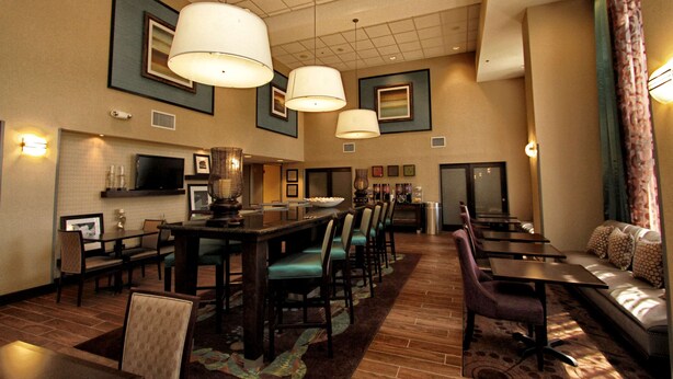 A hotel lobby with a long dining table, multiple chairs, a sofa, and a television on the wall. - Hampton Inn & Suites Bluffton-Sun City (Bluffton)