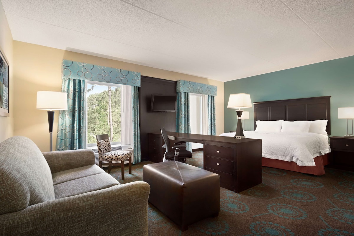 A hotel room with a bed, desk, chair, ottoman, and a television.