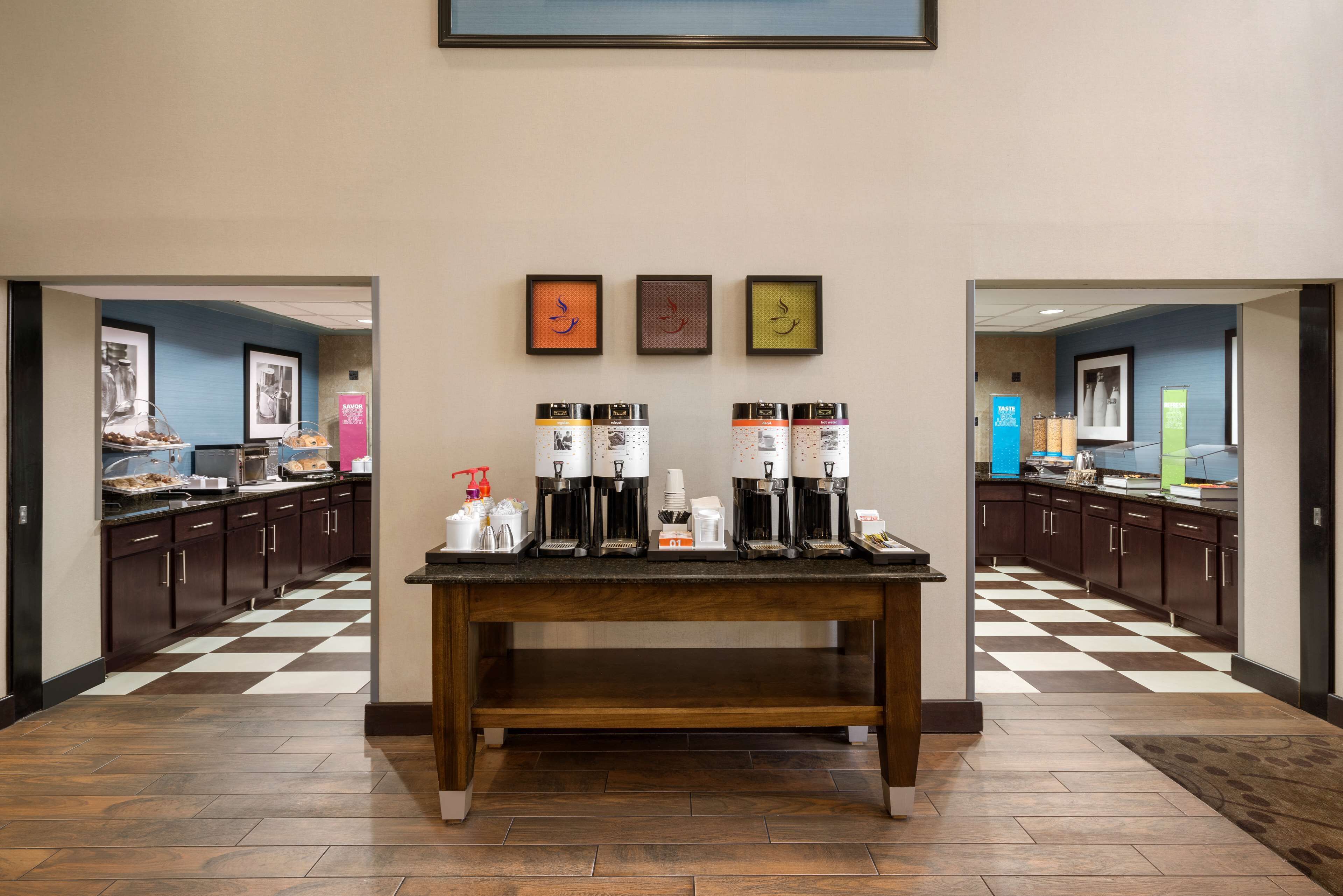 A hotel kitchen with a checkered floor, a coffee station with multiple coffee makers, and framed artwork on the wall.