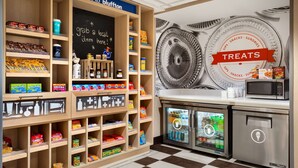 A snack and treat display area with shelves stocked with various snacks, a chalkboard sign, and a refrigerator. - Hampton Inn & Suites Bluffton-Sun City (Bluffton)