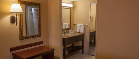 Quarto de hotel com uma cama, um banco de madeira, um banheiro com pia, espelho e porta-toalhas.