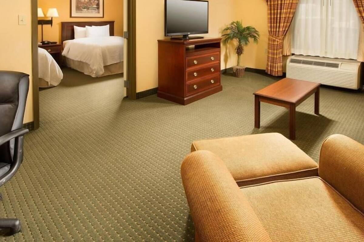 A hotel room with a bed, a television on a wooden dresser, a small table, a chair, and a window with curtains.
