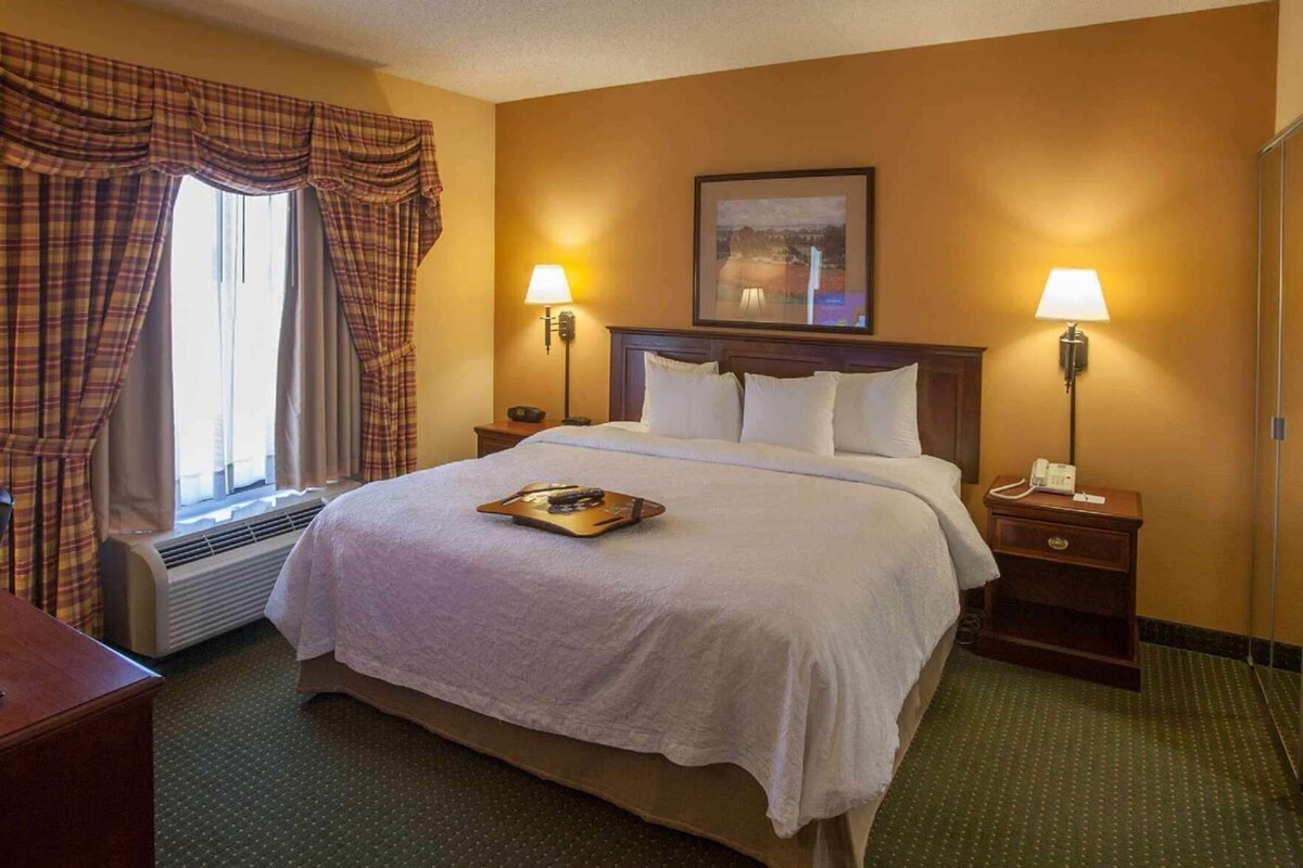 A hotel room with a bed, two bedside lamps, a window with curtains, a painting above the bed, and a bedside table with a phone.