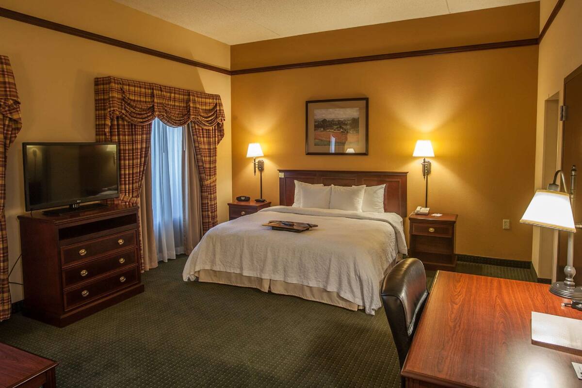 A hotel room with a bed, a TV on a wooden dresser, two bedside lamps, a painting on the wall, and a desk with a lamp.