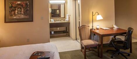 A hotel room with a bed, a desk with a lamp, a chair, a small table with remote controls, a framed picture on the wall, and a bathroom visible through an open door.