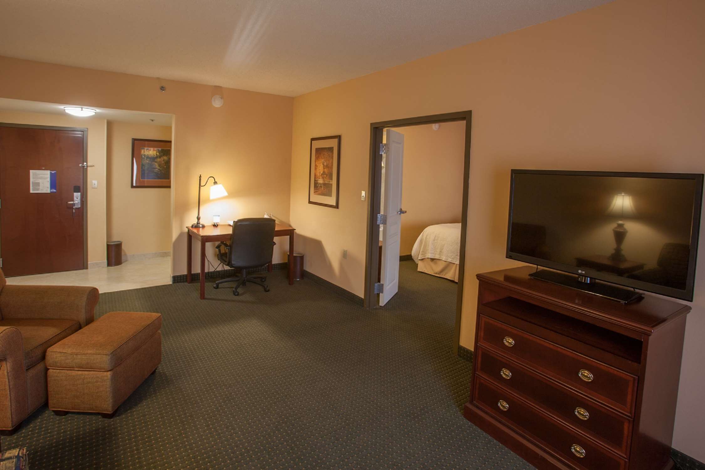 A hotel room with a sofa, a desk with a lamp, a television on a wooden dresser, and a door leading to another room.