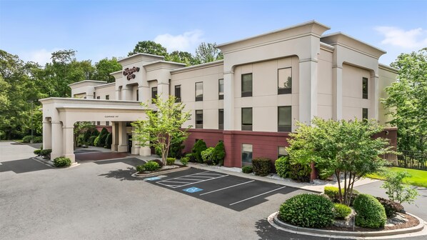 A hotel entrance with a clear sign, a parking area, and landscaped greenery.