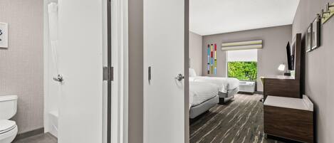 Una habitación de hotel moderna con baño, una cama, una mesita de noche y un ventanal con vistas a zonas verdes.