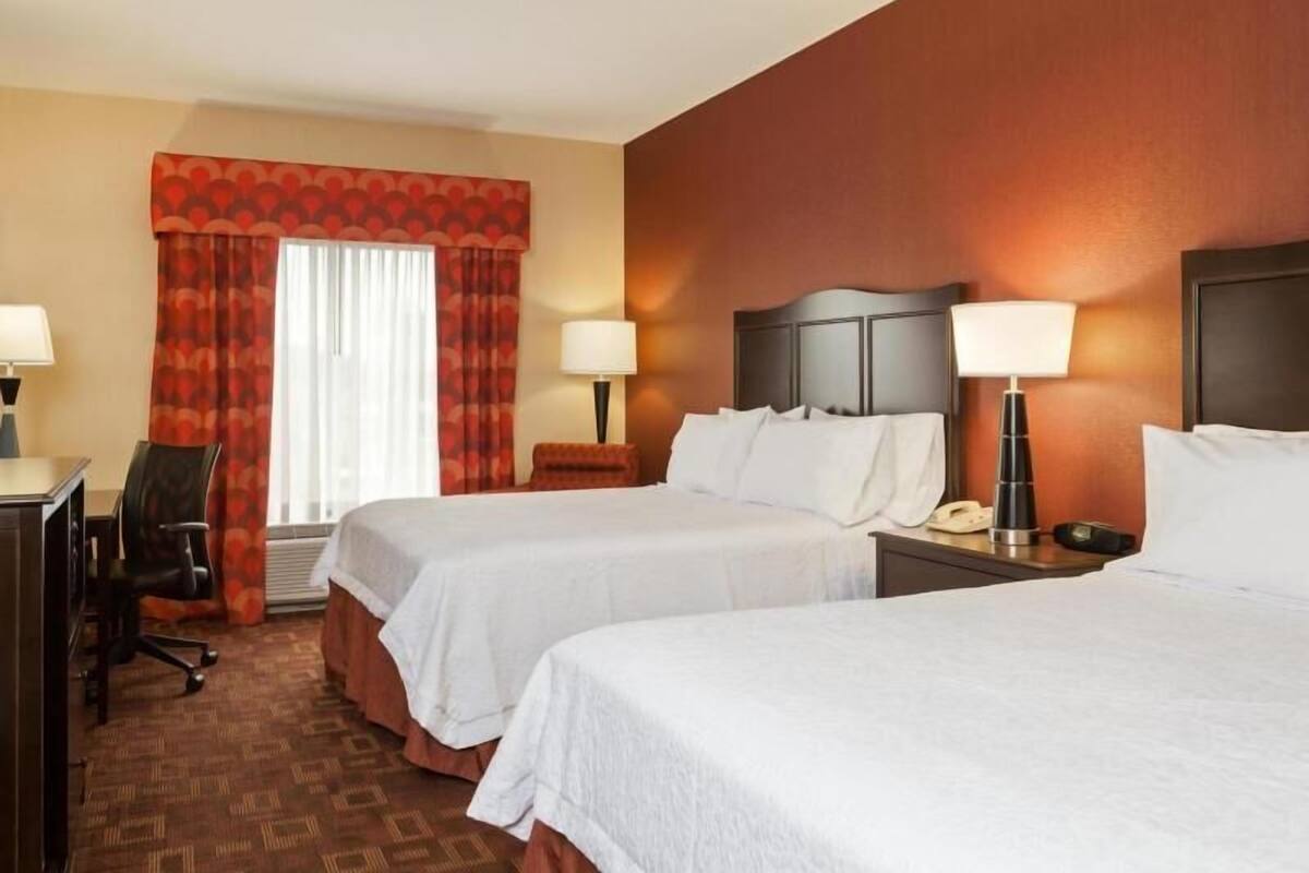 A hotel room with two beds, a desk, a chair, a telephone, and a lamp.