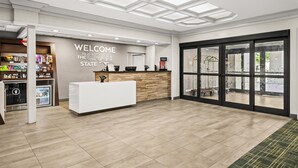 A modern reception area with a reception desk, a welcome sign, a refrigerator with beverages, and a view through large glass doors. - Hampton Inn Elkton (Elkton)