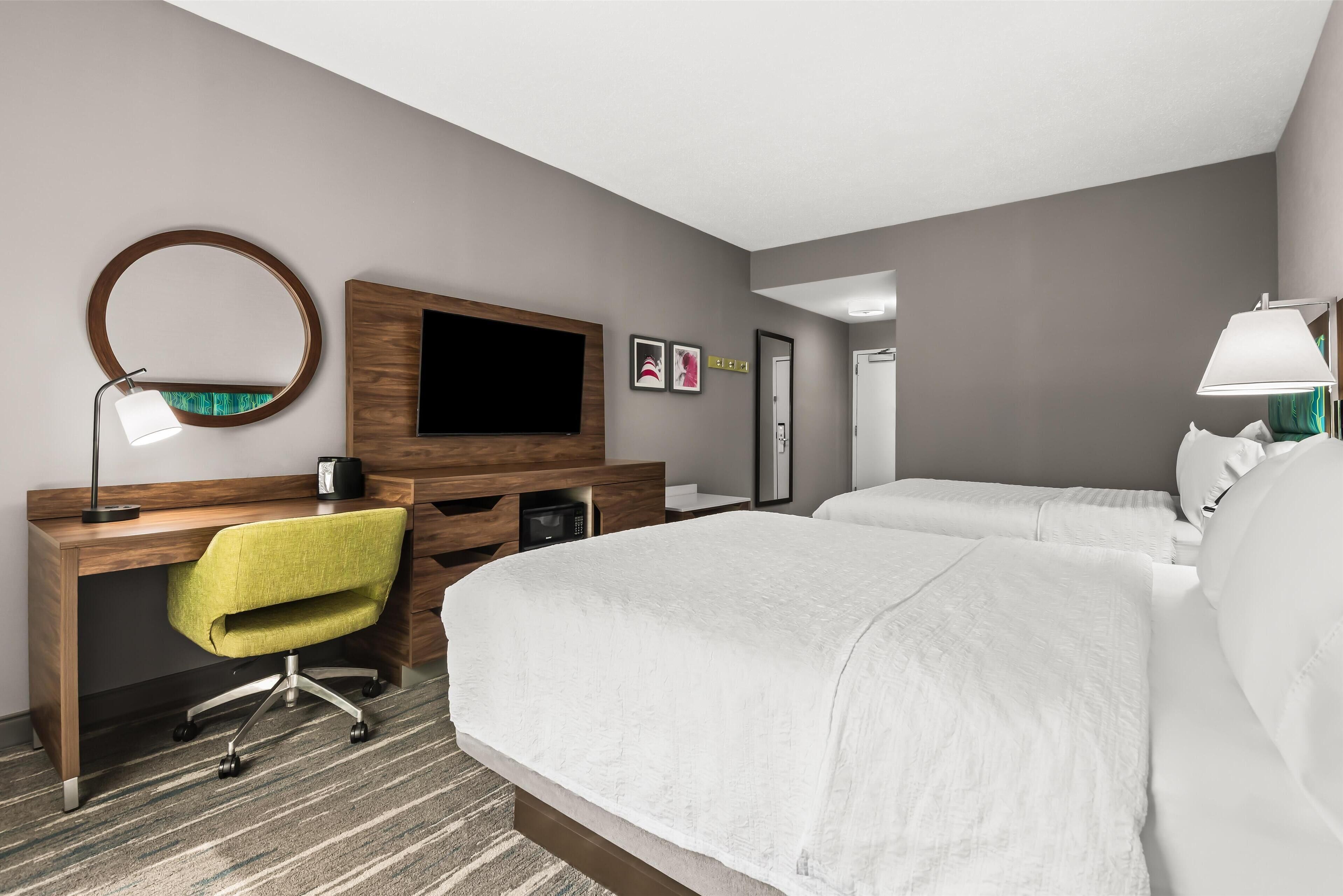 A hotel room with a wooden desk, a green chair, a bed with white bedding, a round mirror, a flat-screen TV, and a bedside lamp.