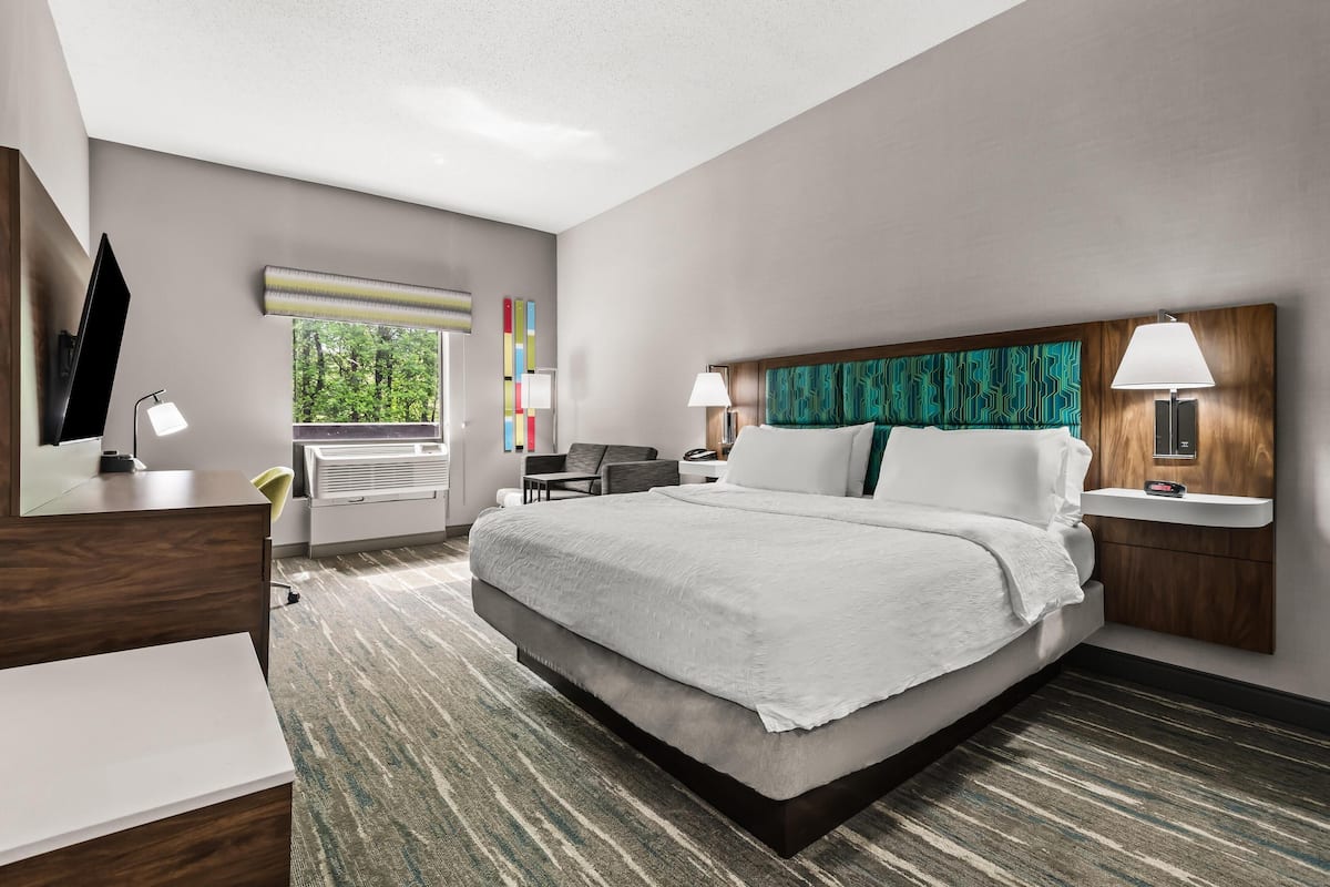 A modern hotel room with a large bed, a desk, a chair, and a window with blinds.