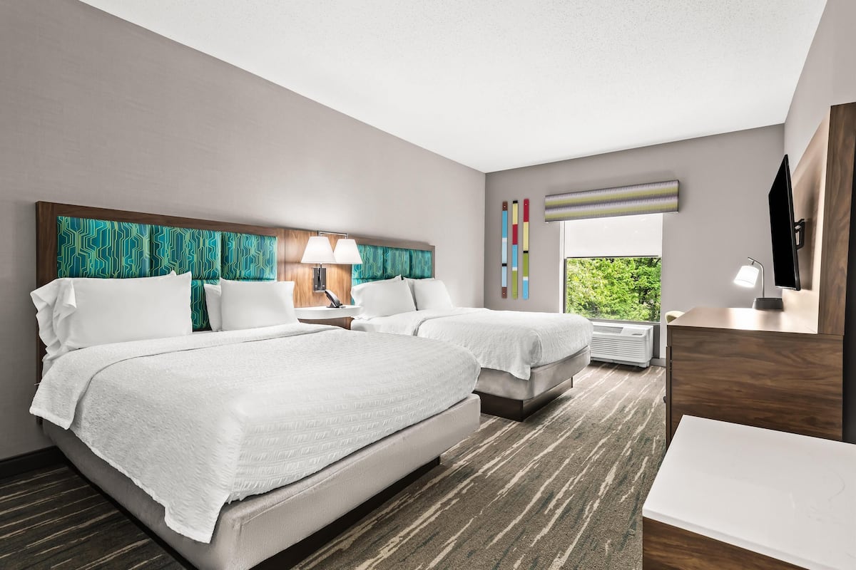 A hotel room with two beds, a desk, a chair, a TV, and a window with a view of greenery.