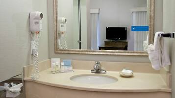 A hotel bathroom with a sink, a mirror, a hairdryer, and a television in the background.