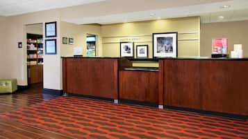 Un área de recepción con un mostrador de madera, una alfombra estampada de color rojo, una pared con fotografías enmarcadas y un cartel de bienvenida, además de una máquina expendedora.