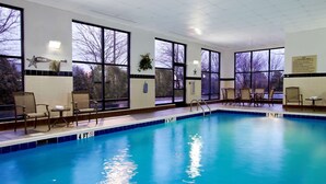 Una piscina interior con agua azul cristalina, rodeada de amplias ventanas y sillas.