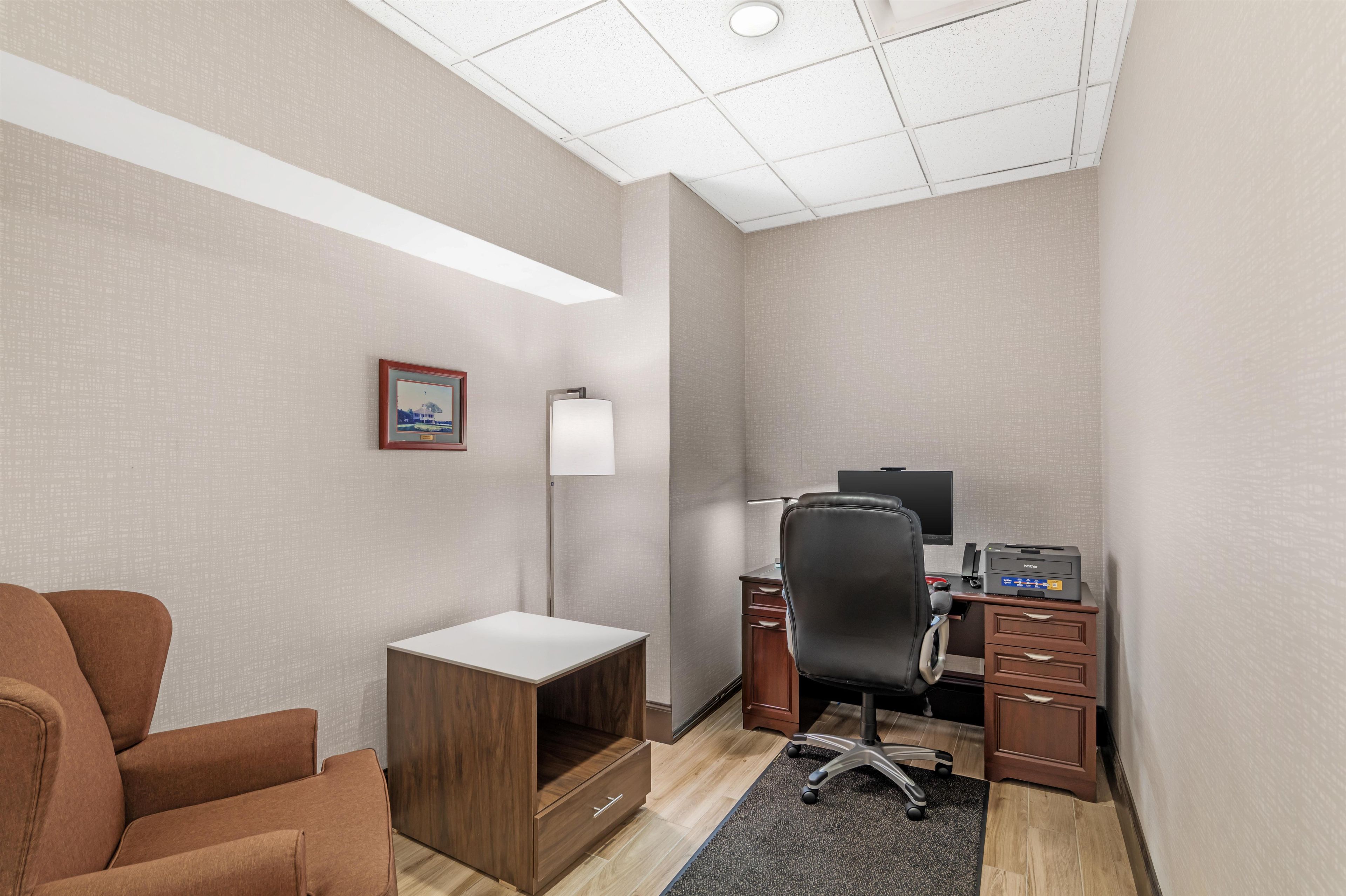 A hotel office room with a desk, chair, lamp, and a framed picture on the wall.