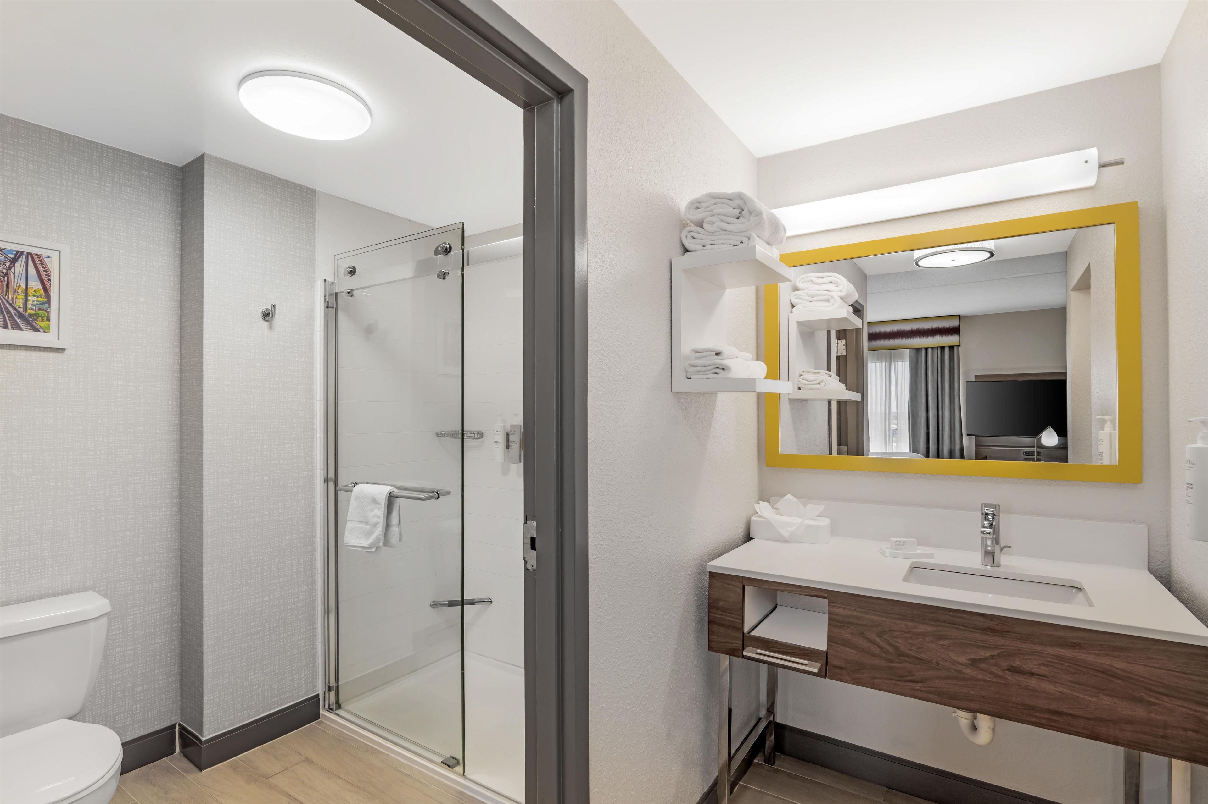 Un baño moderno con ducha envuelta en vidrio, un lavabo blanco con tocador de madera y un espejo con marco amarillo.