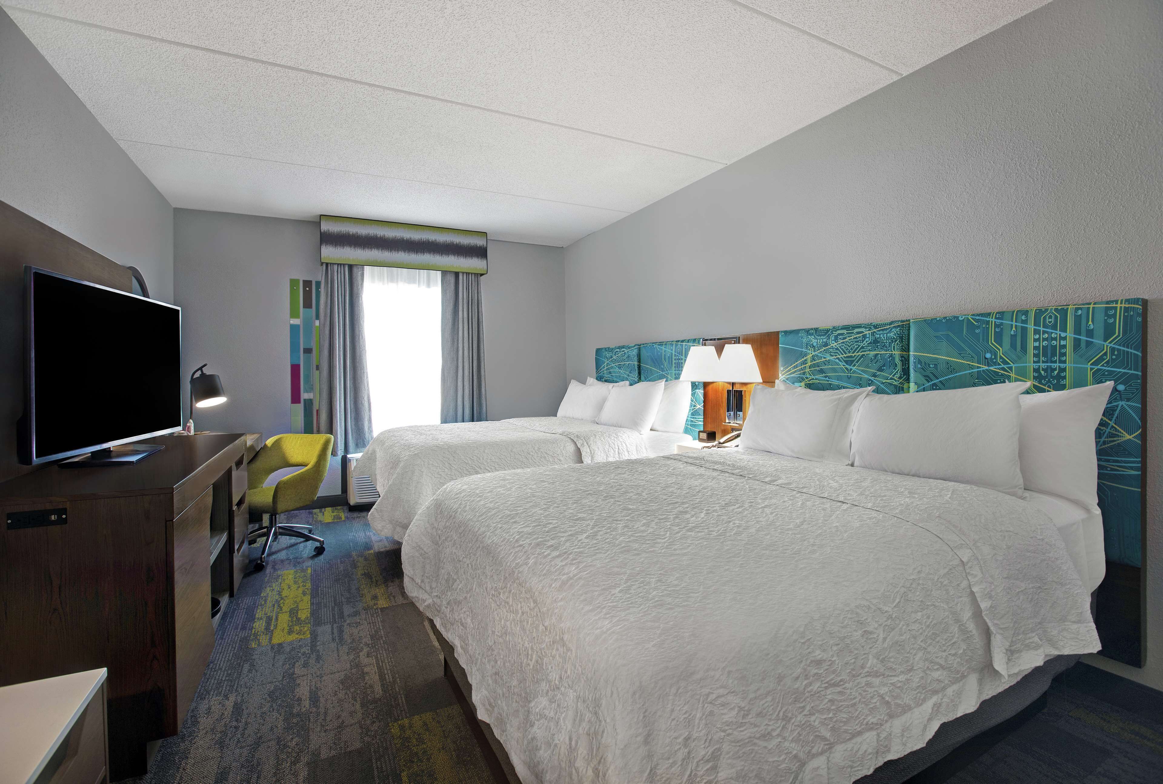 A hotel room with two beds, a desk with a TV, a chair, and a window with curtains.