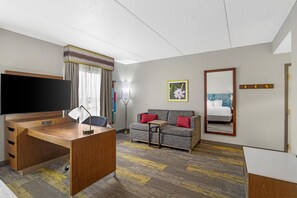 A hotel room with a wooden desk, a flat-screen TV, a sofa with red cushions, a standing lamp, a framed picture on the wall, and a mirror reflecting the bed. - Hampton Inn & Suites Augusta West (Augusta)