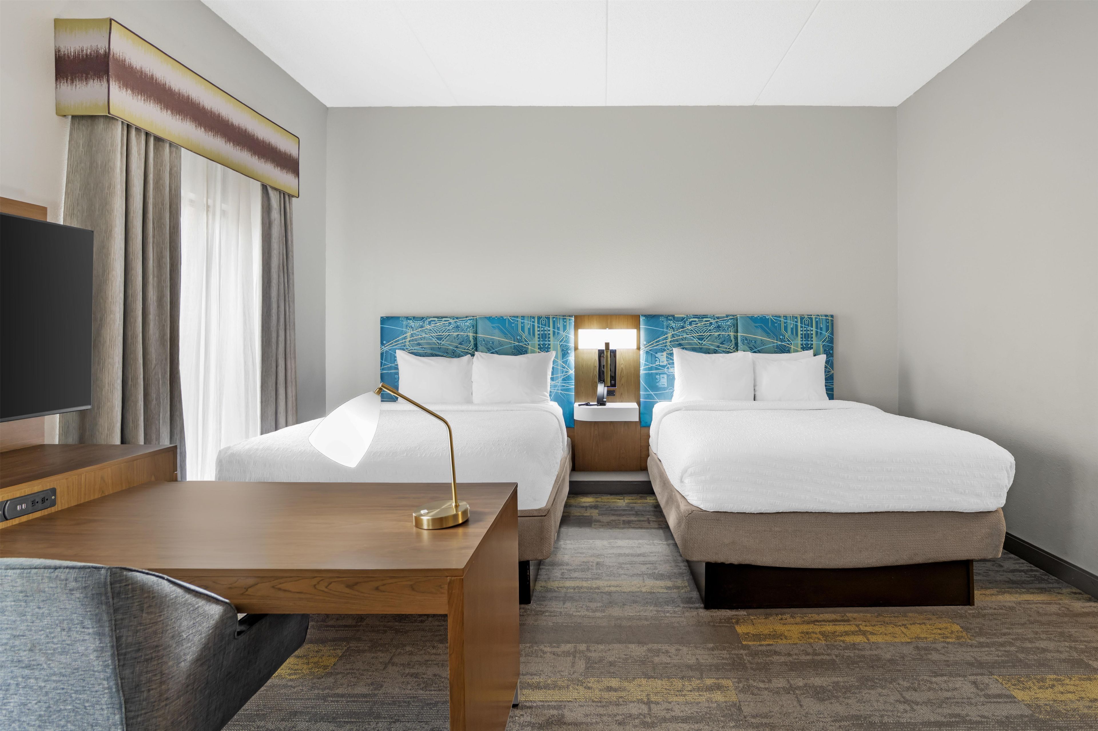 A hotel room with two beds, a desk, a TV, and a large window with curtains.