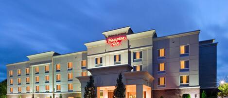 Un hotel Hampton Inn con diseño moderno, que destaca un letrero llamativo, varios pisos y una entrada bien iluminada.