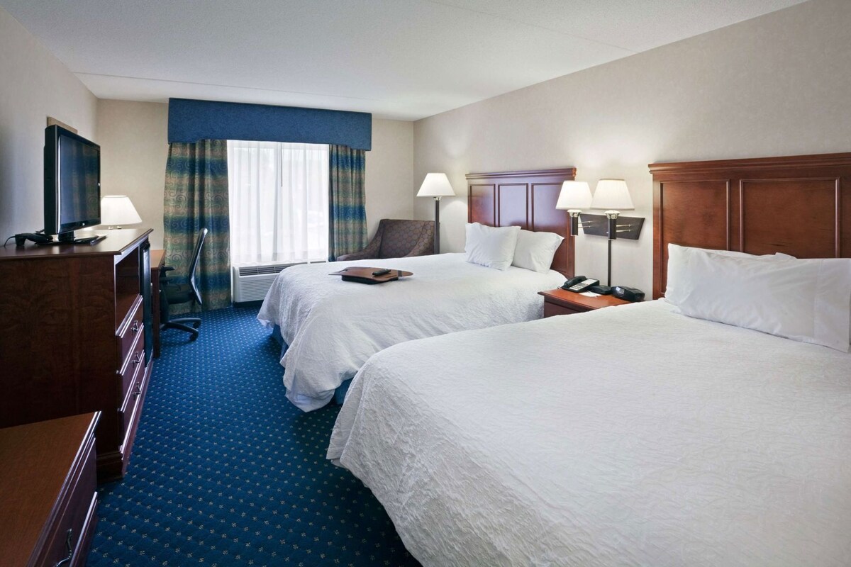 A hotel room with two beds, a desk with a lamp, a chair, and a television.