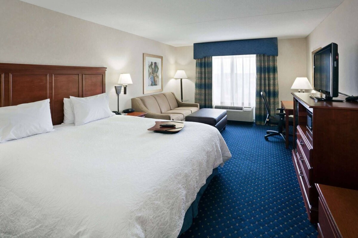 A hotel room with a large bed, a desk, a chair, a television, and a window with curtains.