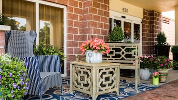 A brick building with a number '620' above the entrance. There is a blue and white patterned chair, a wooden bench, and a small table with a vase of pink flowers.