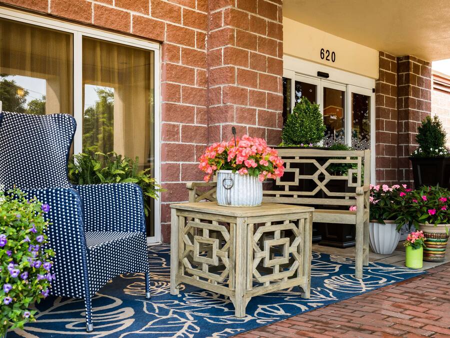 Un edificio de ladrillo con el número '620' sobre la entrada. Hay una silla de color azul y blanco con un diseño, un banco de madera y una pequeña mesa con un jarrón de flores rosas.