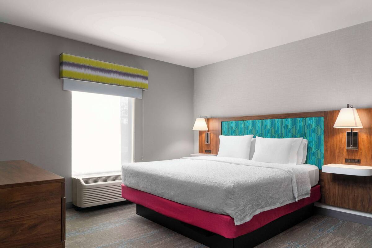 A hotel room with a bed, a nightstand, a lamp, a window with blinds, and a wall-mounted air conditioner.