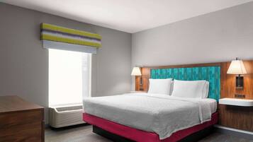 A hotel room with a bed, a nightstand, a lamp, a window with blinds, and a wall-mounted air conditioner.