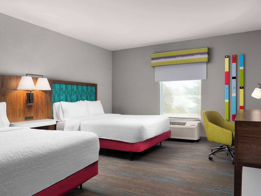 A hotel room with two beds, a desk, a chair, a window with blinds, a wall-mounted lamp, and a colorful abstract painting.