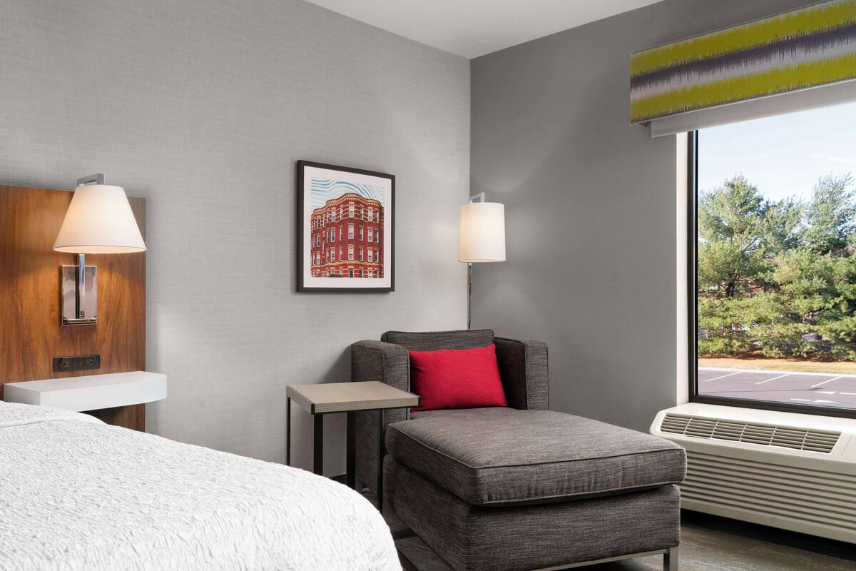 A hotel room with a bed, a gray sofa with a red pillow, a small table, a wall-mounted lamp, a framed picture, and a window with a view of greenery.