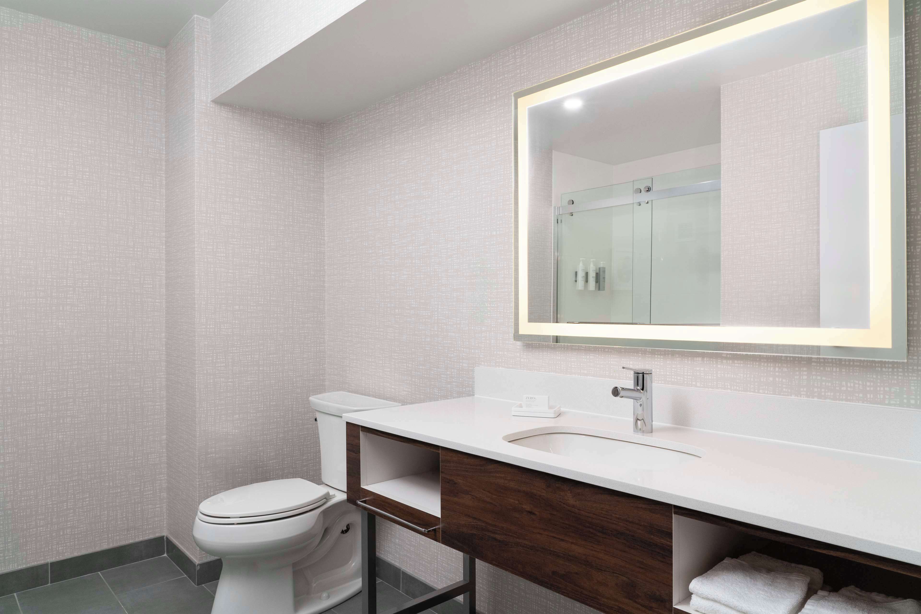 A modern bathroom with a white toilet, a wooden vanity, a large mirror, and a tiled wall.