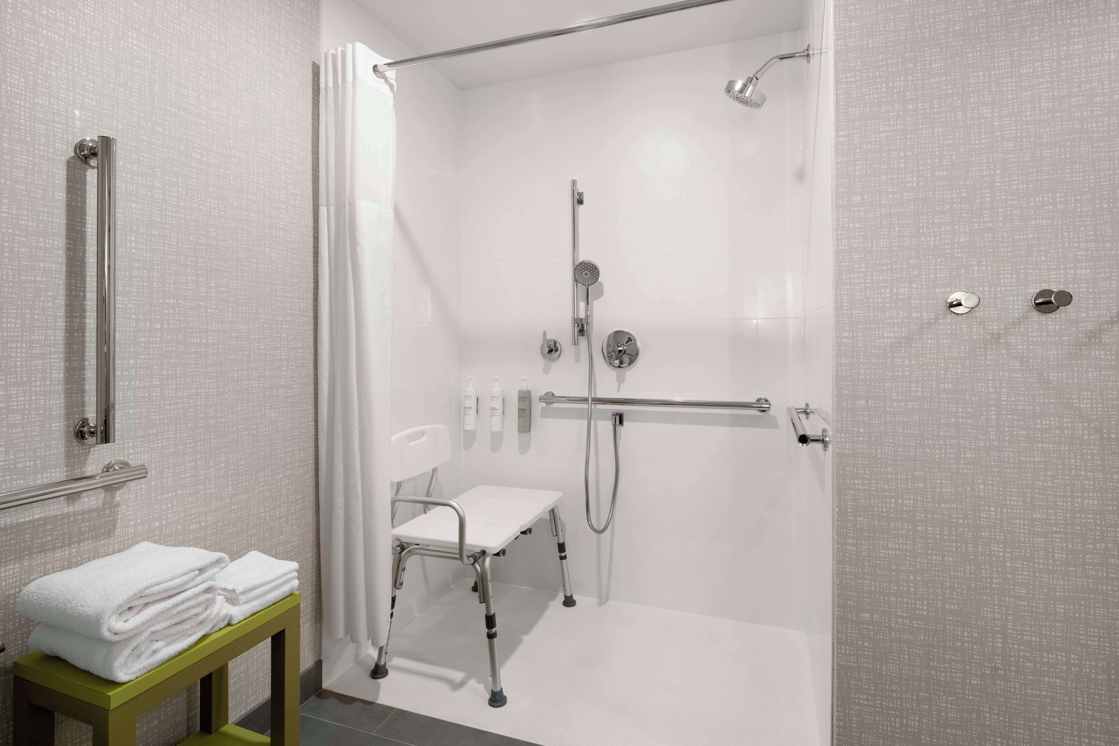 A modern bathroom with a walk-in shower, grab bars, a wheelchair-accessible bench, and neatly stacked towels.