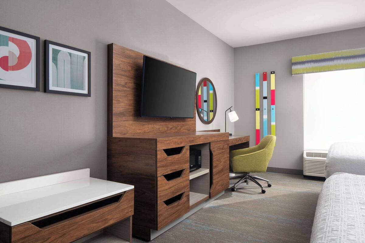 A modern hotel room with a flat-screen TV mounted on a wooden cabinet, a desk with drawers, a green chair, and a bed with a white comforter.