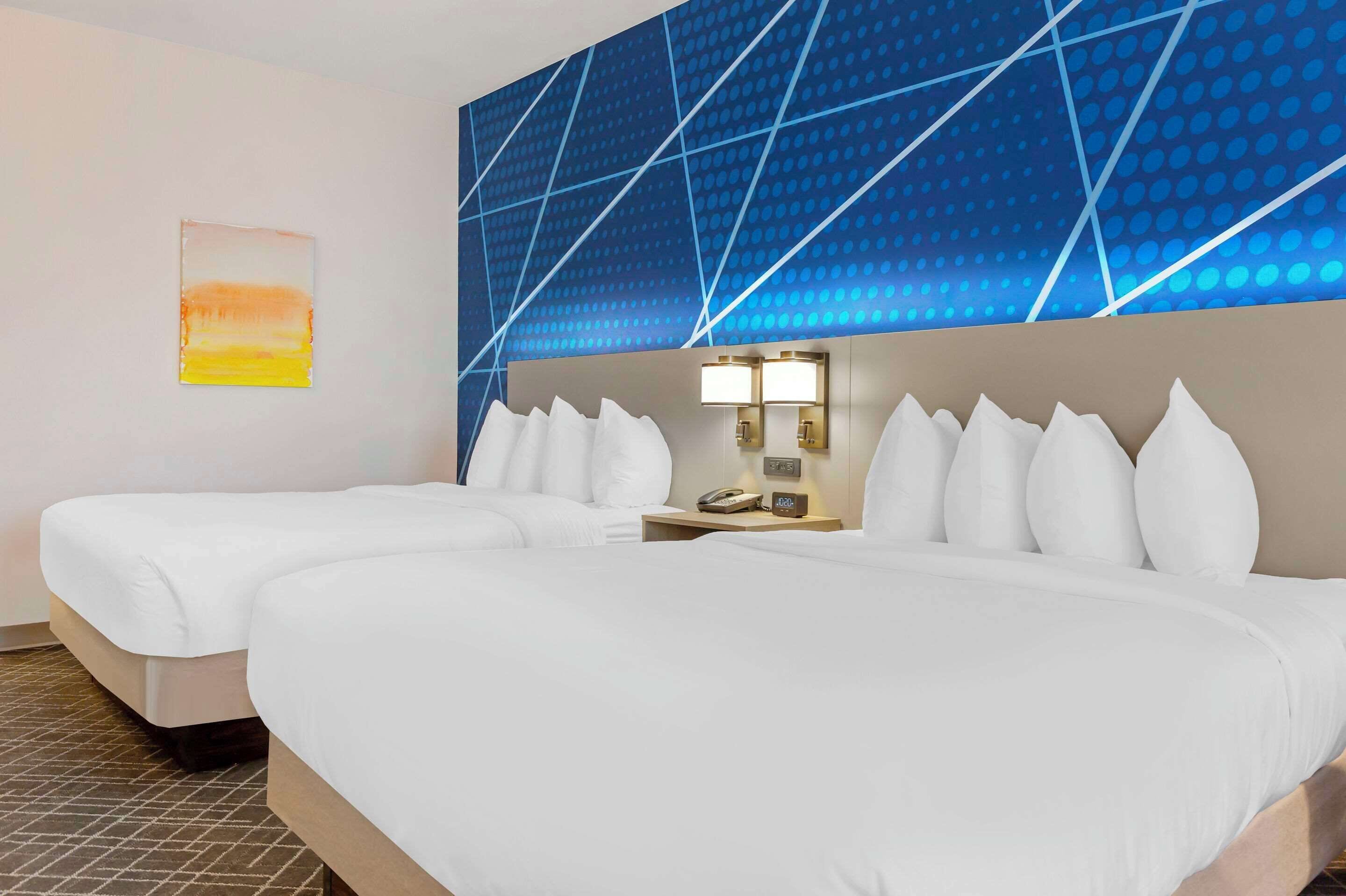 Habitación de hotel con dos camas, una mesita de noche con lámpara y una pared con diseño geométrico.