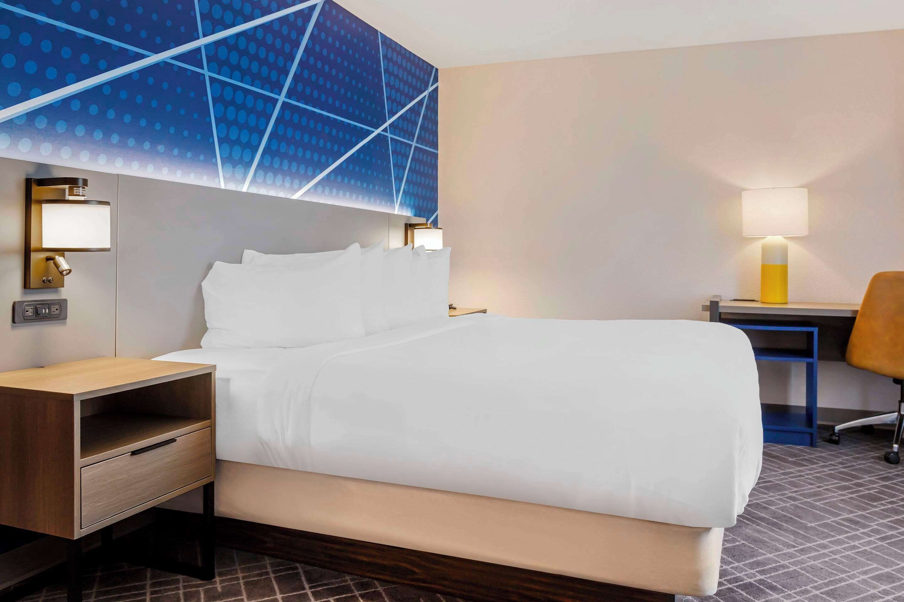 Une chambre d’hôtel équipée d’un lit, d’un bureau, d’une chaise, d’une table de chevet et d’une lampe fixée au mur.
