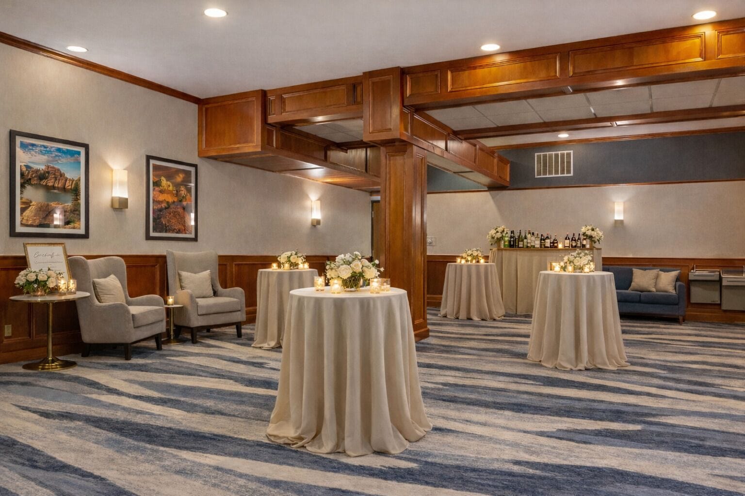 A hotel room with a large, open layout featuring a high ceiling, wooden paneling, and a carpeted floor. There are several round tables with candles and flowers, and a bar area with bottles and glasses.