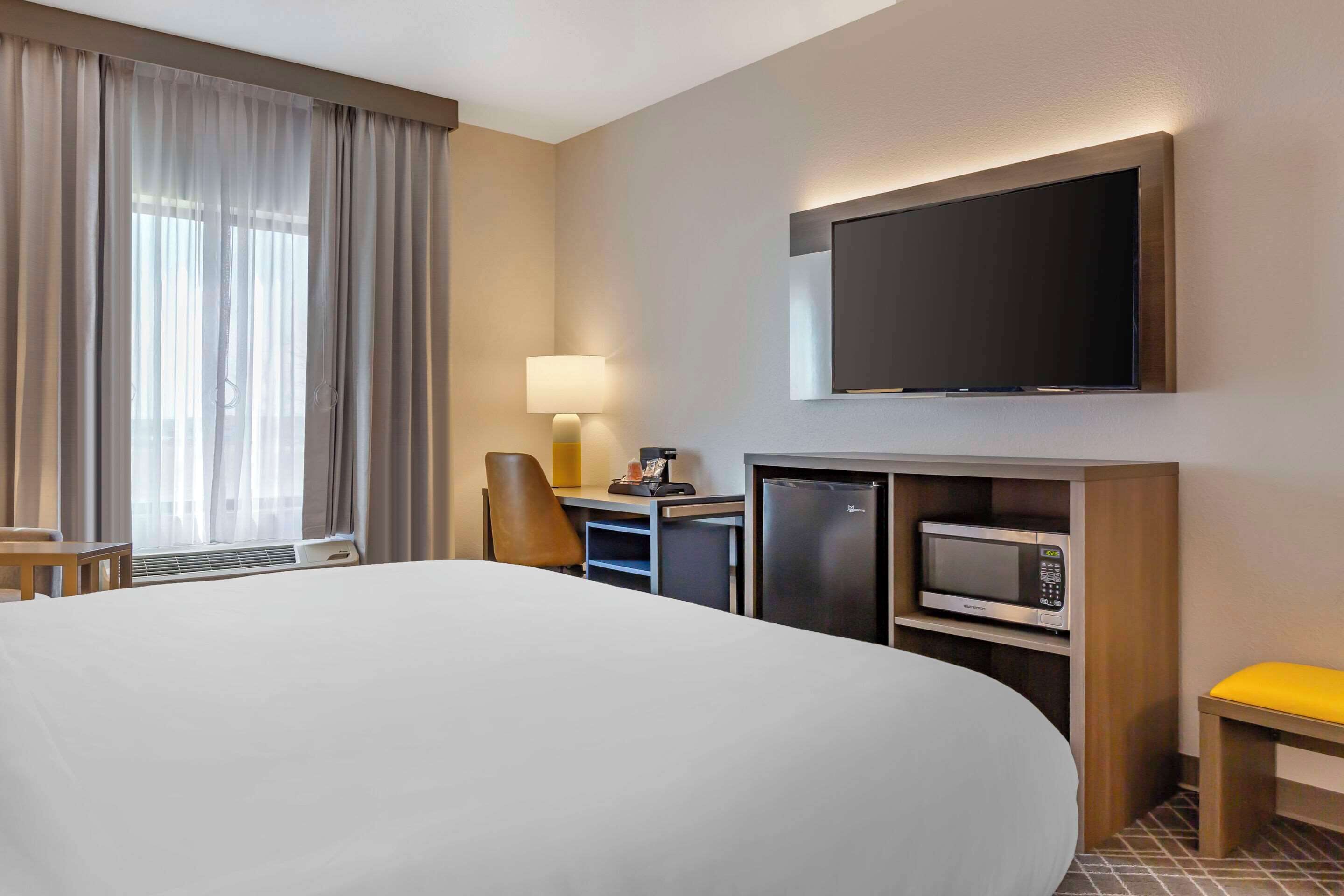 A hotel room with a large bed, a desk with a computer, a microwave, and a flat-screen TV mounted on the wall.