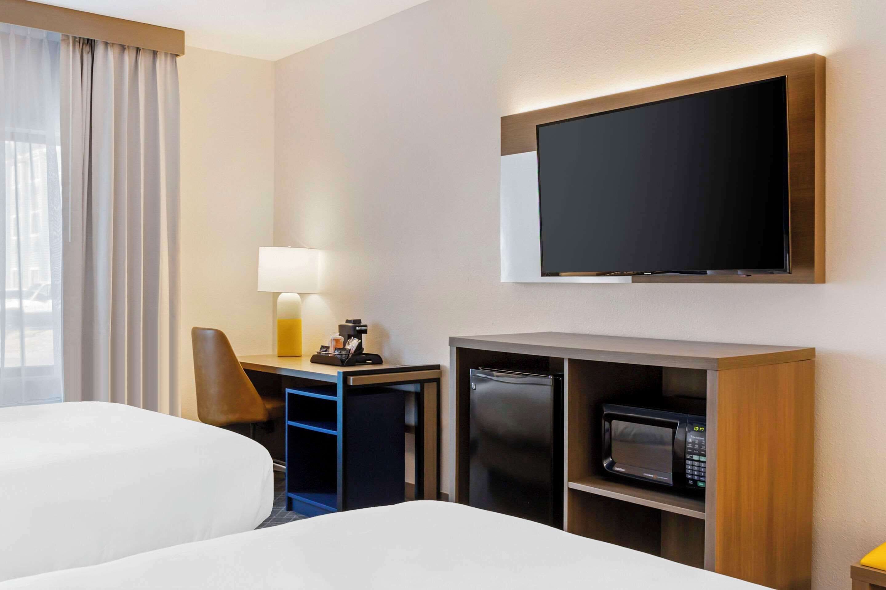 Quarto de hotel com duas camas, uma mesa com luminária, televisão na parede e micro-ondas em armário.