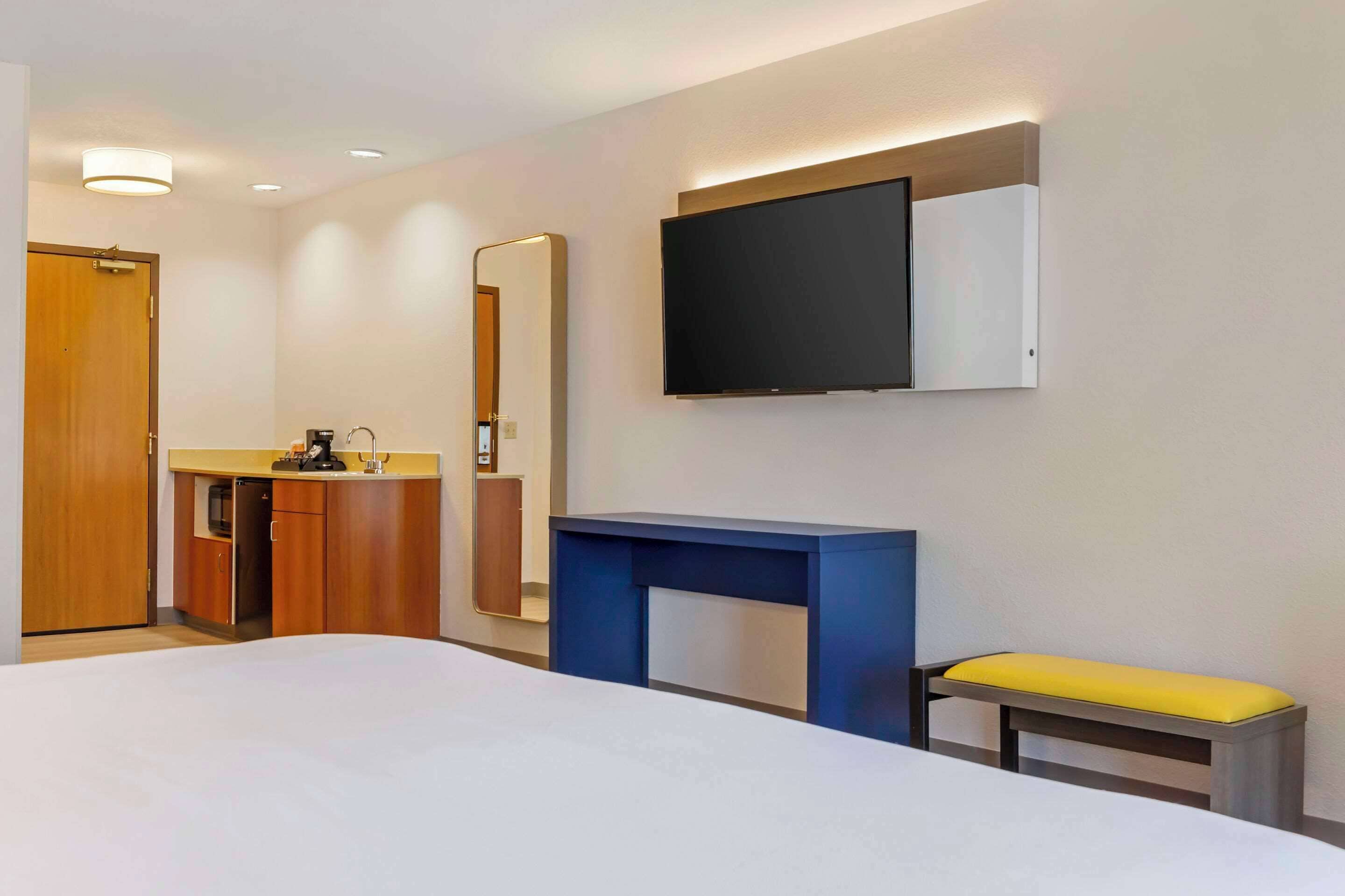 A hotel room with a bed, a desk, a mirror, a television, and a small bench.